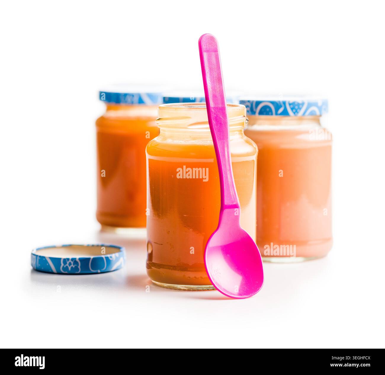 Tre vasetti in vetro di purea arancione con coperchi blu e un cucchiaio rosa brillante, decorati su sfondo bianco. Alimentazione sana fatta in casa, snack per bambini Foto Stock