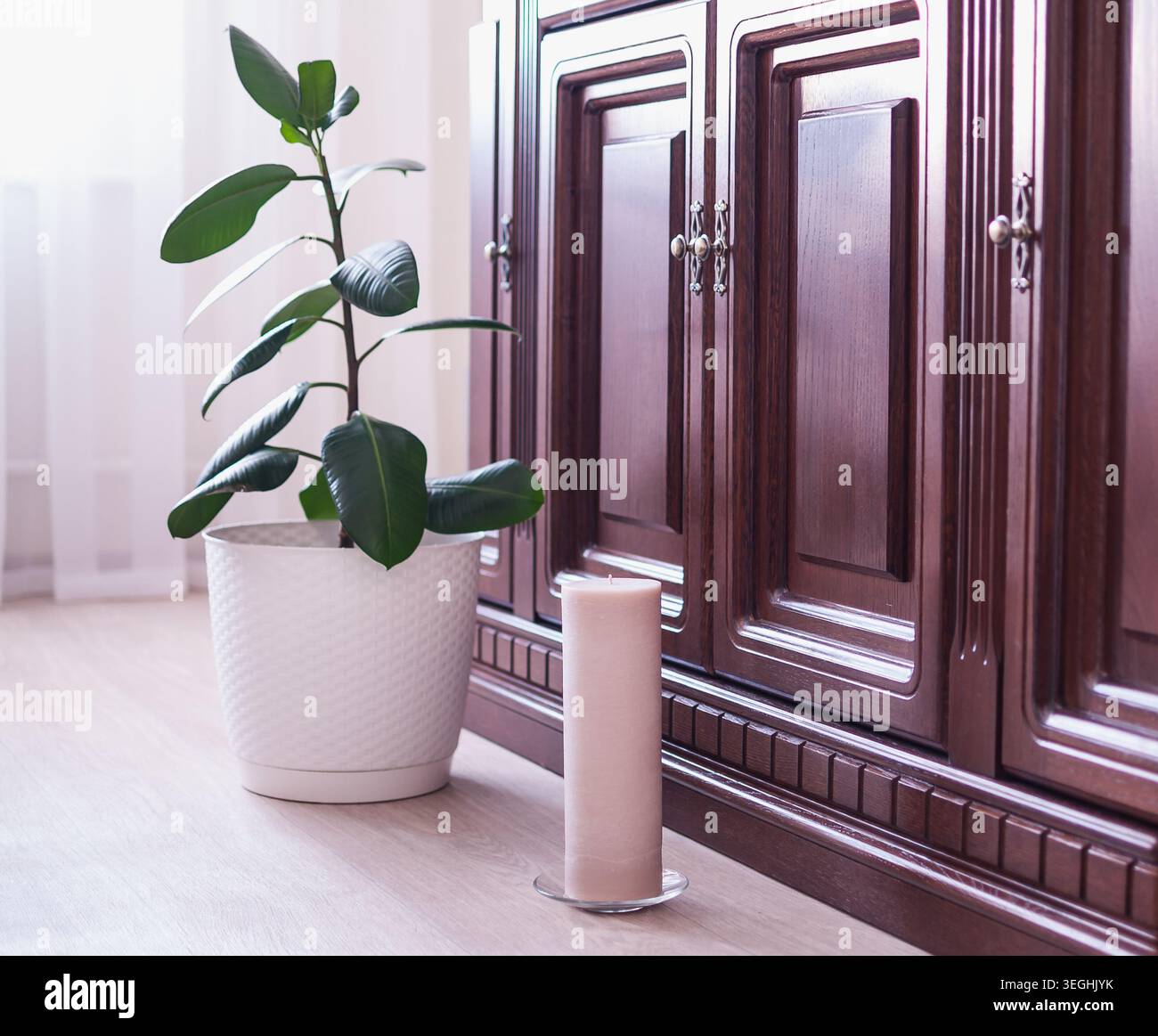 Una grande candela beige siede al piano accanto ad una pianta verde interna in una pentola bianca vicino ad un classico armadietto di legno scuro Foto Stock