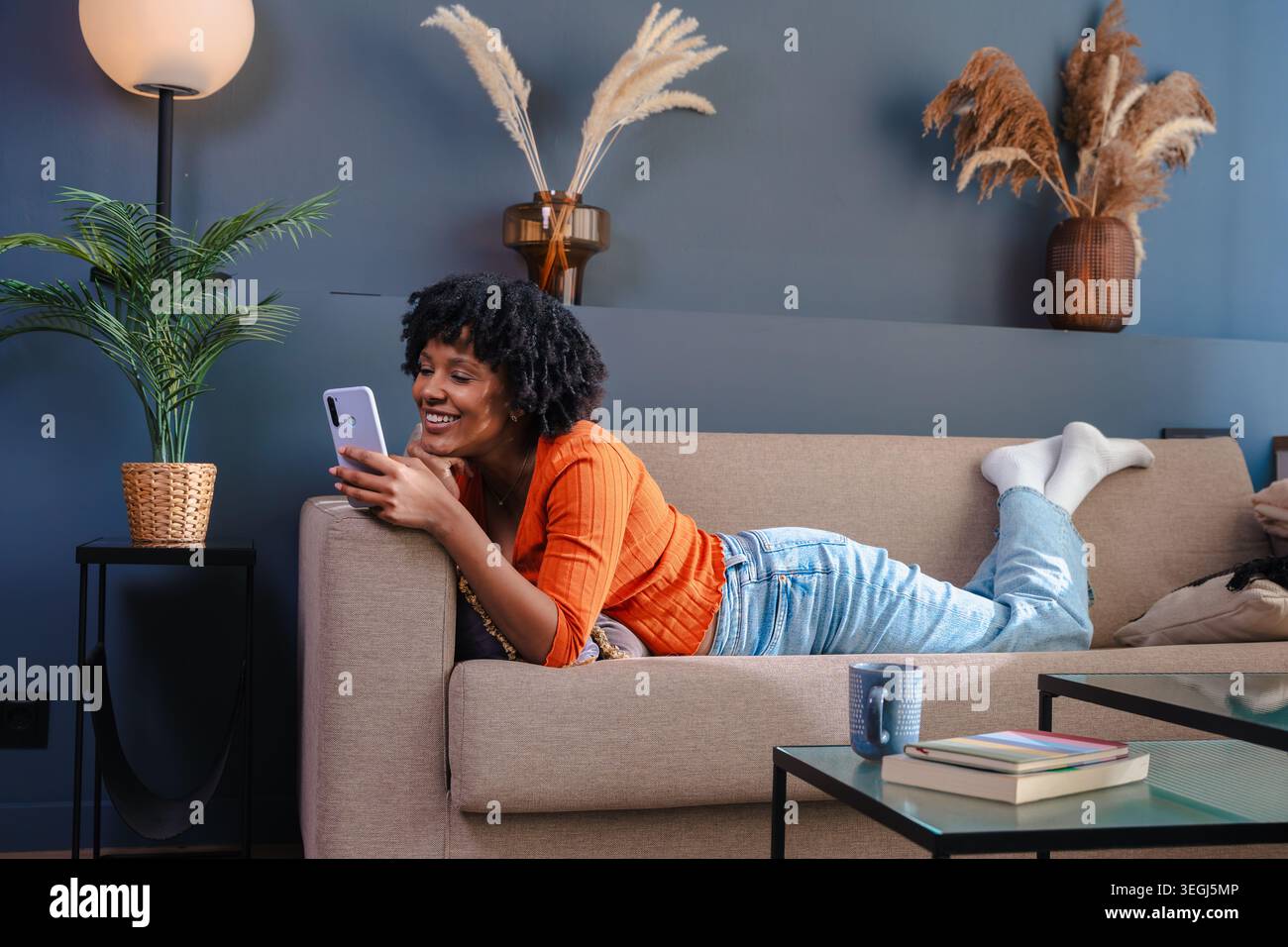 Donna afroamericana sdraiata sul divano e che usa il suo smartphone a casa Foto Stock