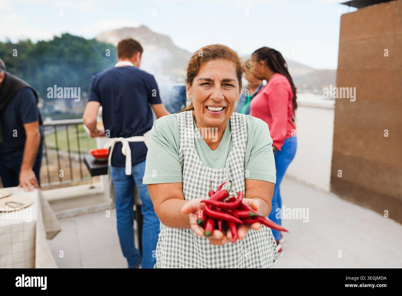 Donna latina anziana che tiene in mano il peperoncino fresco mentre cucina il pranzo al barbecue sullo sfondo - persone multigenerazionali che si divertono insieme durante il fine settimana Foto Stock