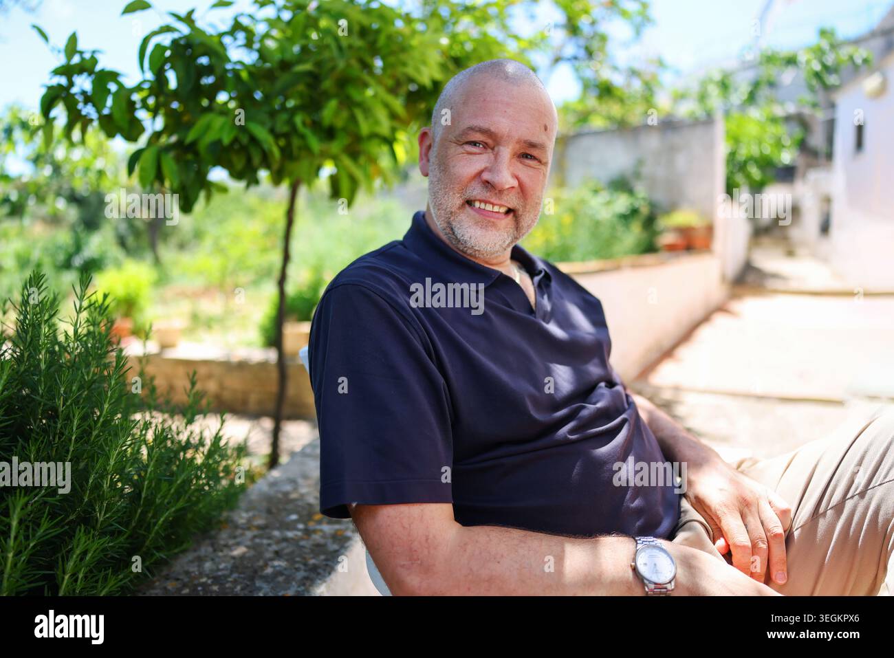 Uomo maturo sorridente che si rilassa all'aperto nel soleggiato giardino mediterraneo, godendo di una vacanza tranquilla e di un'autentica atmosfera di viaggio estiva. Foto Stock