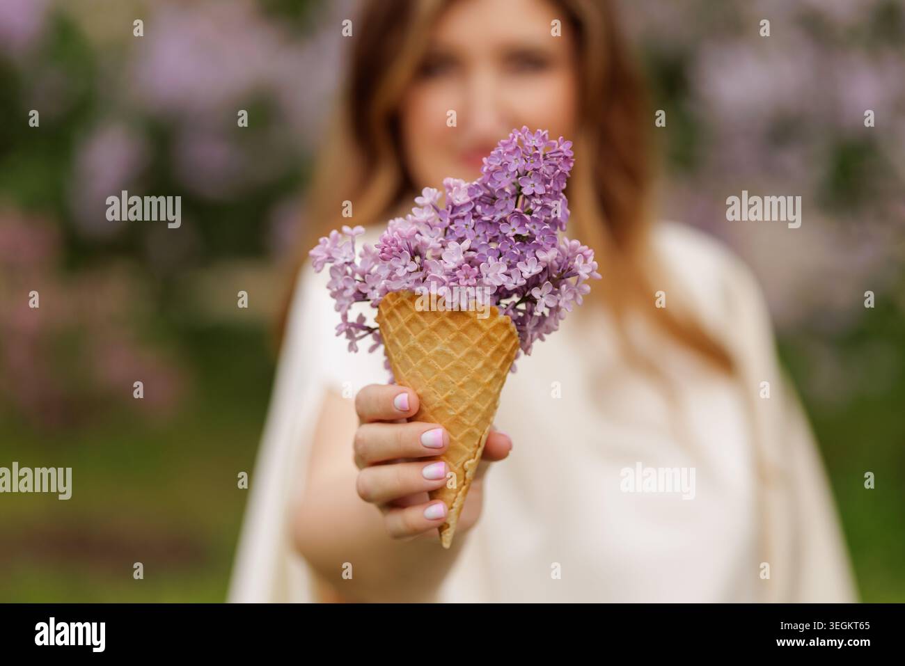 Una donna presenta con gioia un cono di gelato pieno di vivaci fiori lilla, perfetto per il marketing a tema primaverile, composizioni floreali o stile di vita Foto Stock