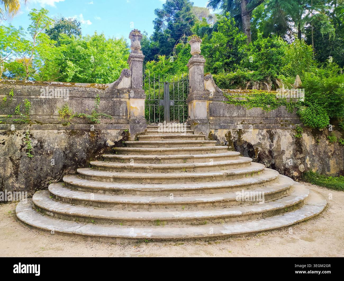 Coimbra, Portogallo - 15 maggio 2026: Storica scalinata semicircolare in pietra che conduce ad un cancello di ferro ornato fiancheggiato da pilastri intemprati in una lussureggiante botanica Foto Stock