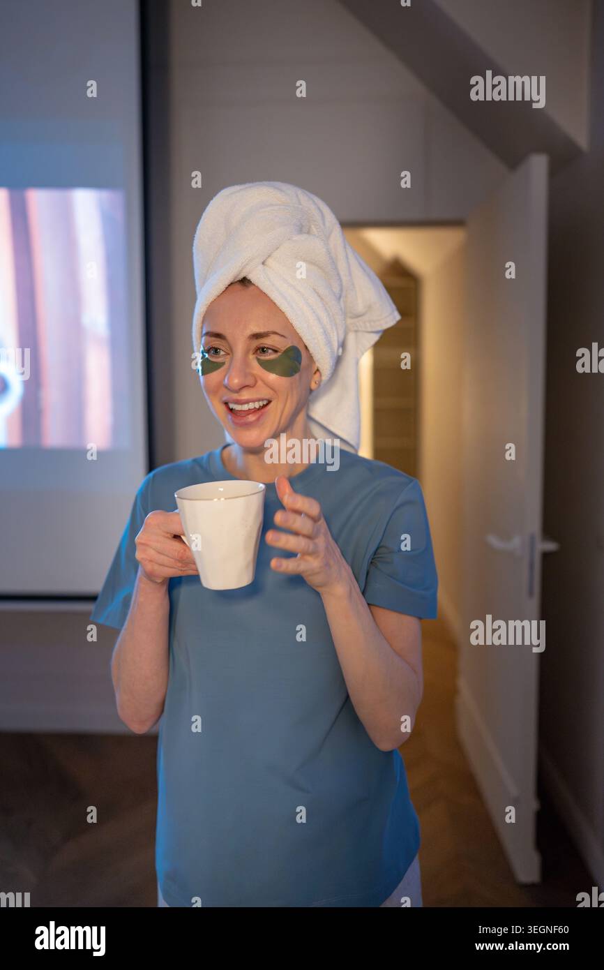 Donna felice sorridente con un asciugamano sulla testa e toppe sotto gli occhi che tengono la tazza godendosi una piacevole serata. Rituale di bellezza self care, relax a casa, Foto Stock