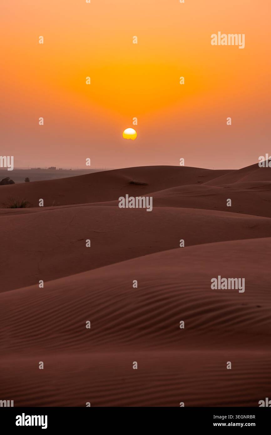 Il sole tramonta su dune di sabbia ondulate in un vasto deserto, creando un caldo bagliore nel cielo. Il paesaggio è tranquillo e vasto, mostrando la bellezza di un tramonto nel paesaggio naturale. Foto Stock
