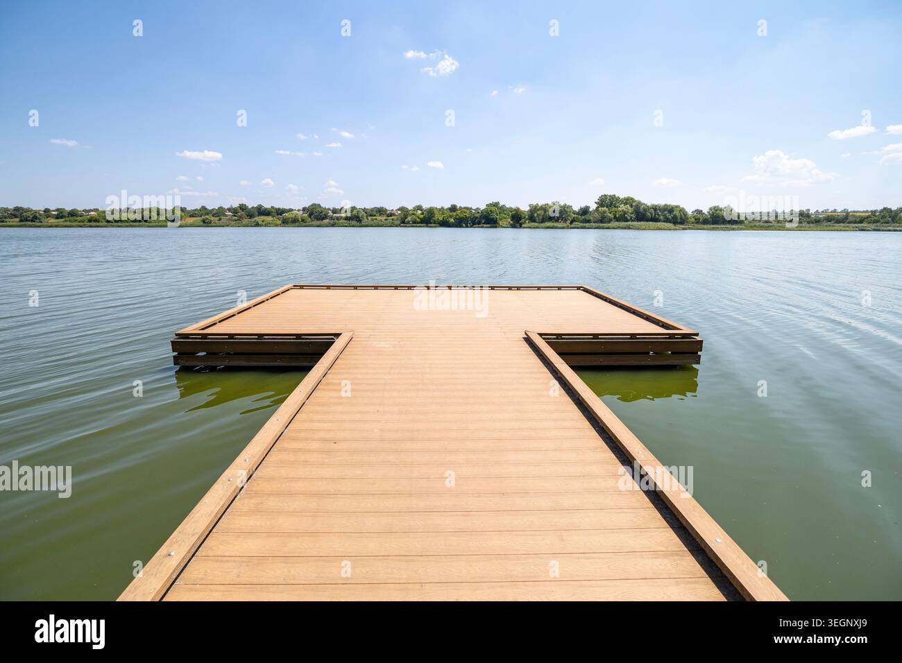 Una banchina di legno vuota a forma di T si estende dal primo piano in un fiume calmo con acqua verde sotto un cielo azzurro limpido con nuvole sparse, con tane Foto Stock
