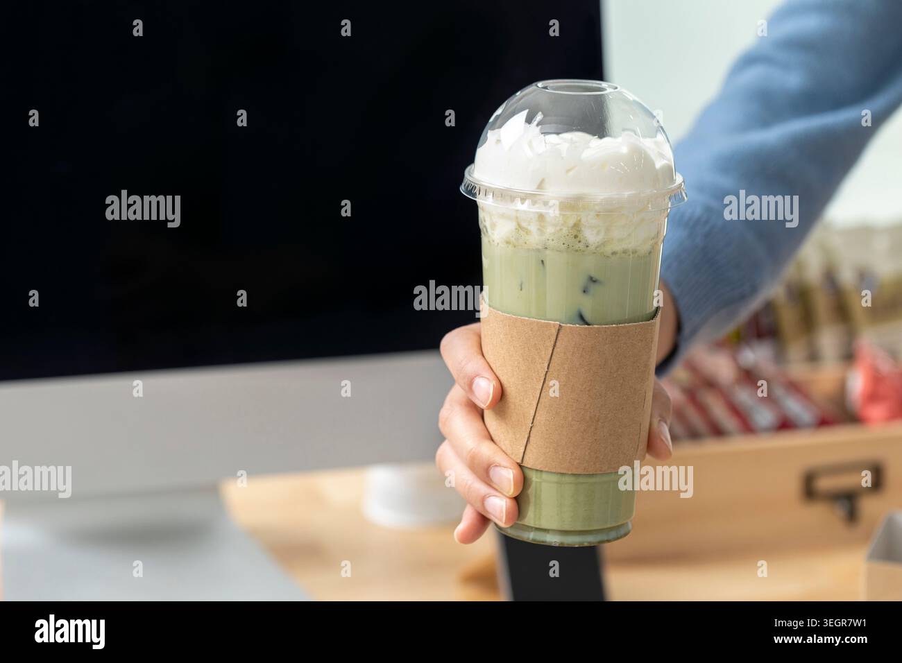 Una persona che tiene in mano un latte matcha alto ghiacciato in una tazza di plastica trasparente con un manicotto di carta, ricoperto di panna montata. Foto Stock