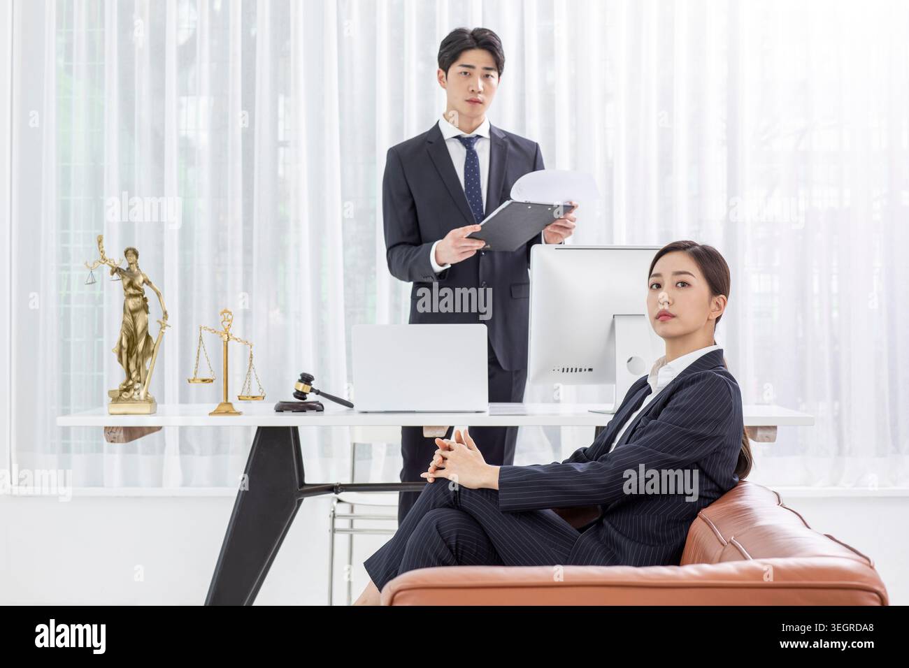 Un giovane uomo d'affari asiatico sta dietro una donna nell'ufficio di un avvocato, rivedendo un fascicolo mentre lei si siede e aspetta. Foto Stock