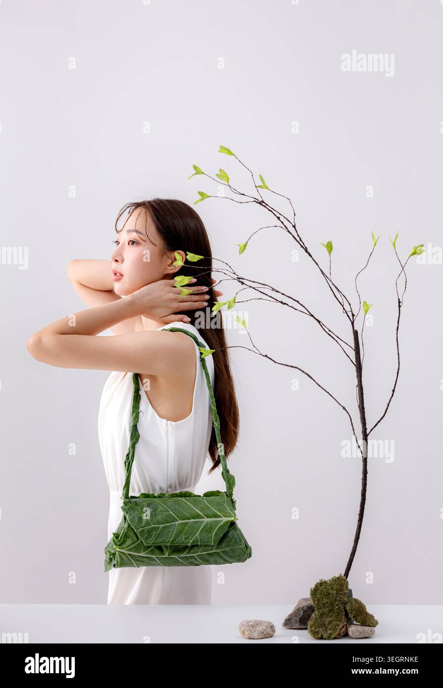 Una donna con un vestito bianco tiene una borsa verde a foglia e tocca i capelli, circondata da un piccolo albero con nuove foglie. Foto Stock