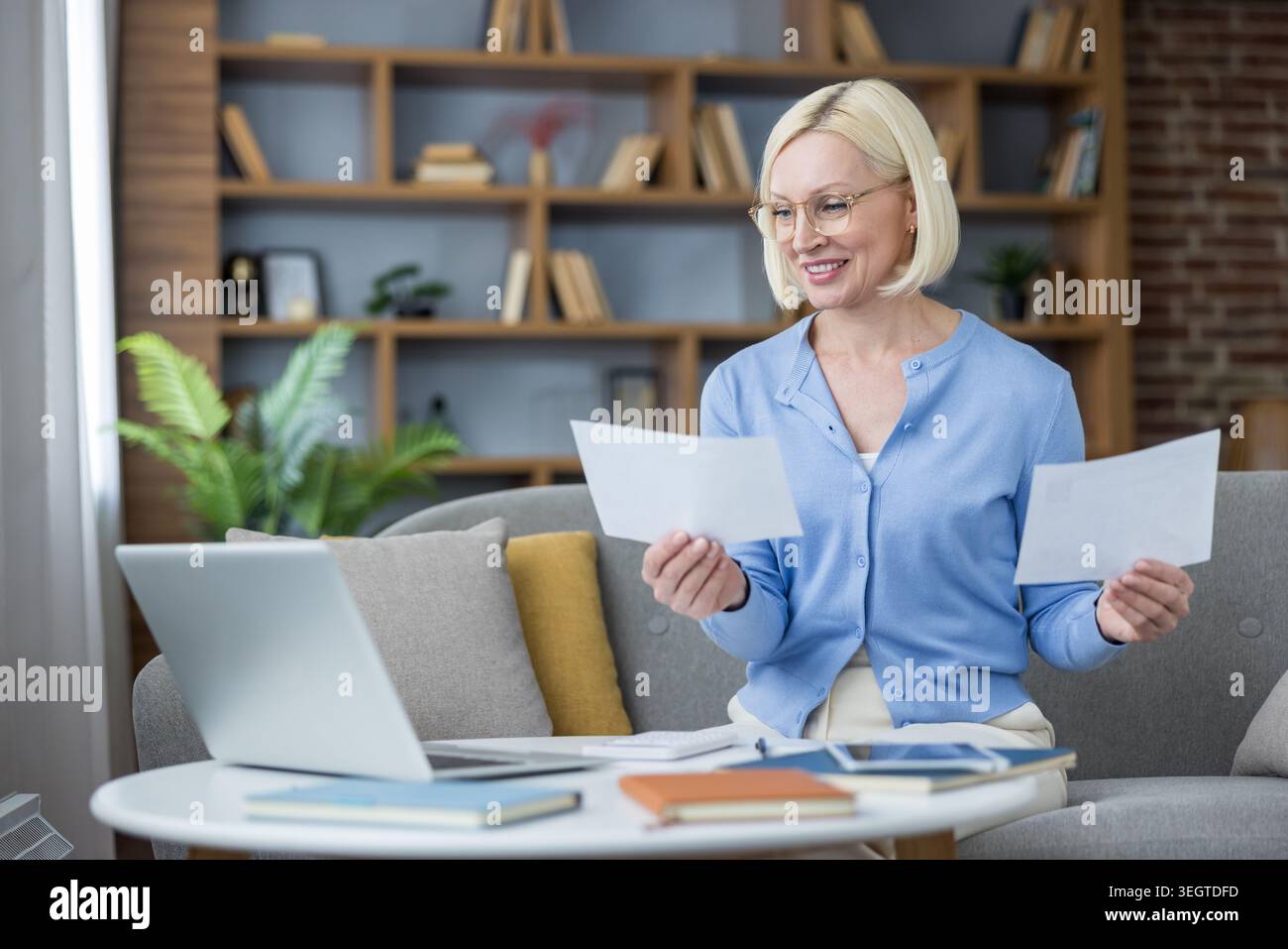 Donna d'affari matura in un accogliente ufficio domestico che esamina documenti e controlla il laptop per la finanza e il lavoro online, sorridendo con fiducia mentre organizza documenti alla sua scrivania Foto Stock