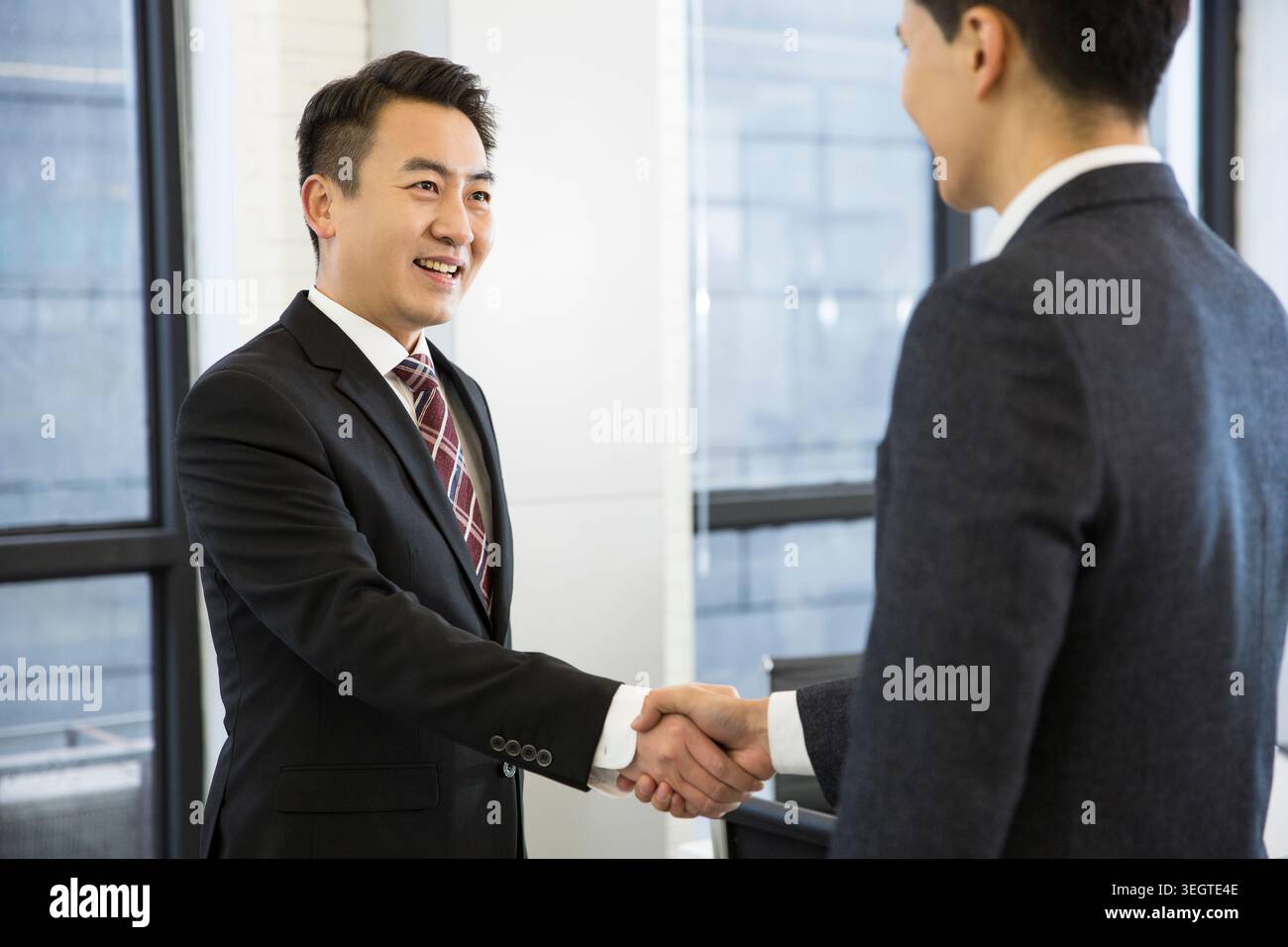 Due uomini d'affari asiatici in tuta che stringono la mano in un ambiente d'ufficio. Foto Stock