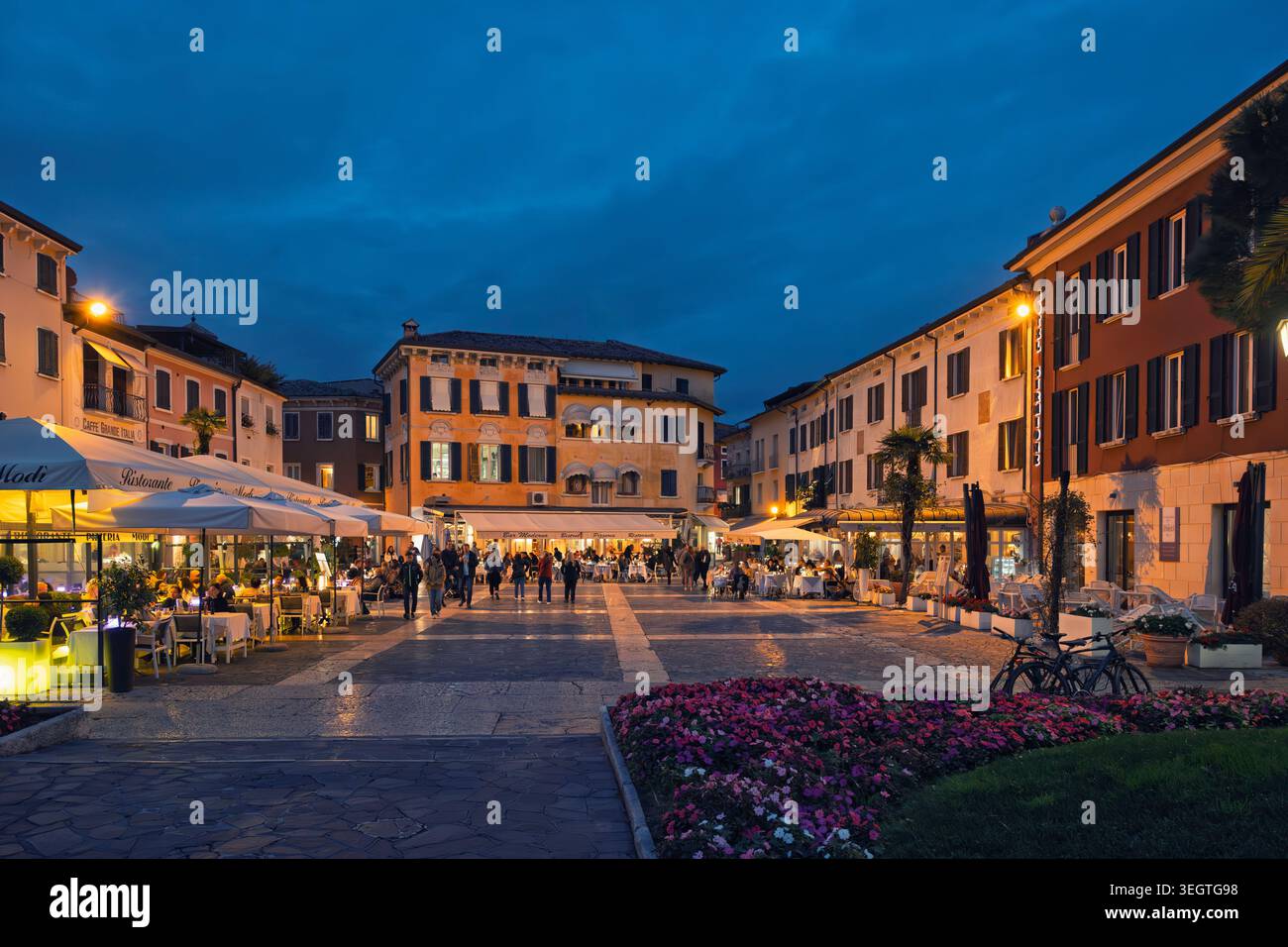 Sirmione, Italia - 04.10.2024: Un'incantevole piazza pedonale al crepuscolo, con edifici storici, caffetterie all'aperto e una calda illuminazione stradale. Foto Stock