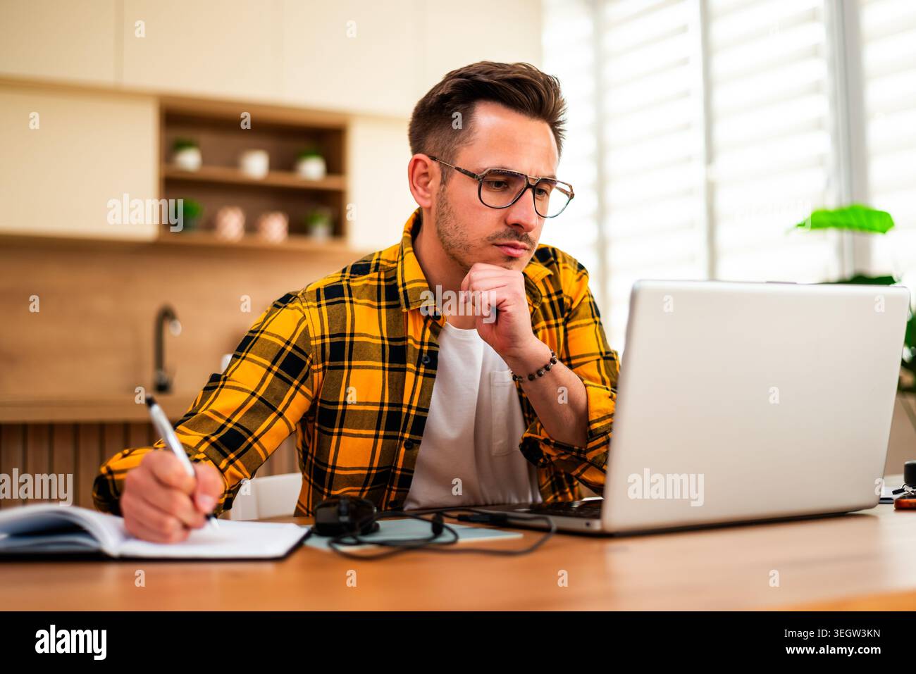 Con un'espressione pensiva, un uomo lavora sul suo portatile e scrive in blocco note, sembrando analizzare le informazioni o risolvere un problema online. Foto Stock