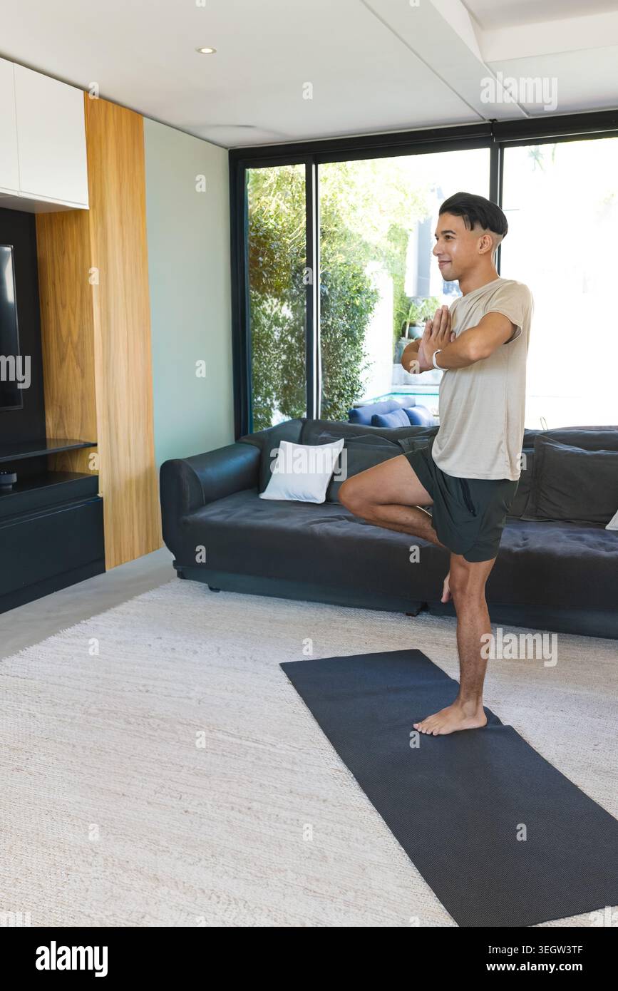 Uomo asiatico che pratica posare gli alberi a piedi nudi su un tappeto da yoga nero nel moderno soggiorno vicino alle finestre Foto Stock