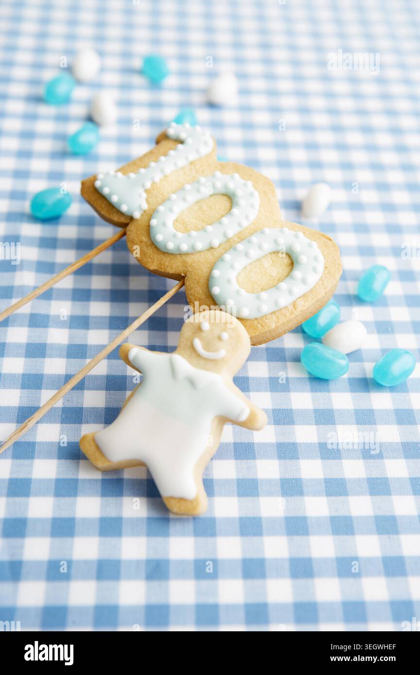Un biscotto sorridente si distingue da un lotto di biscotti a forma di numero 100 su una tovaglia a scacchi blu e bianco. Foto Stock