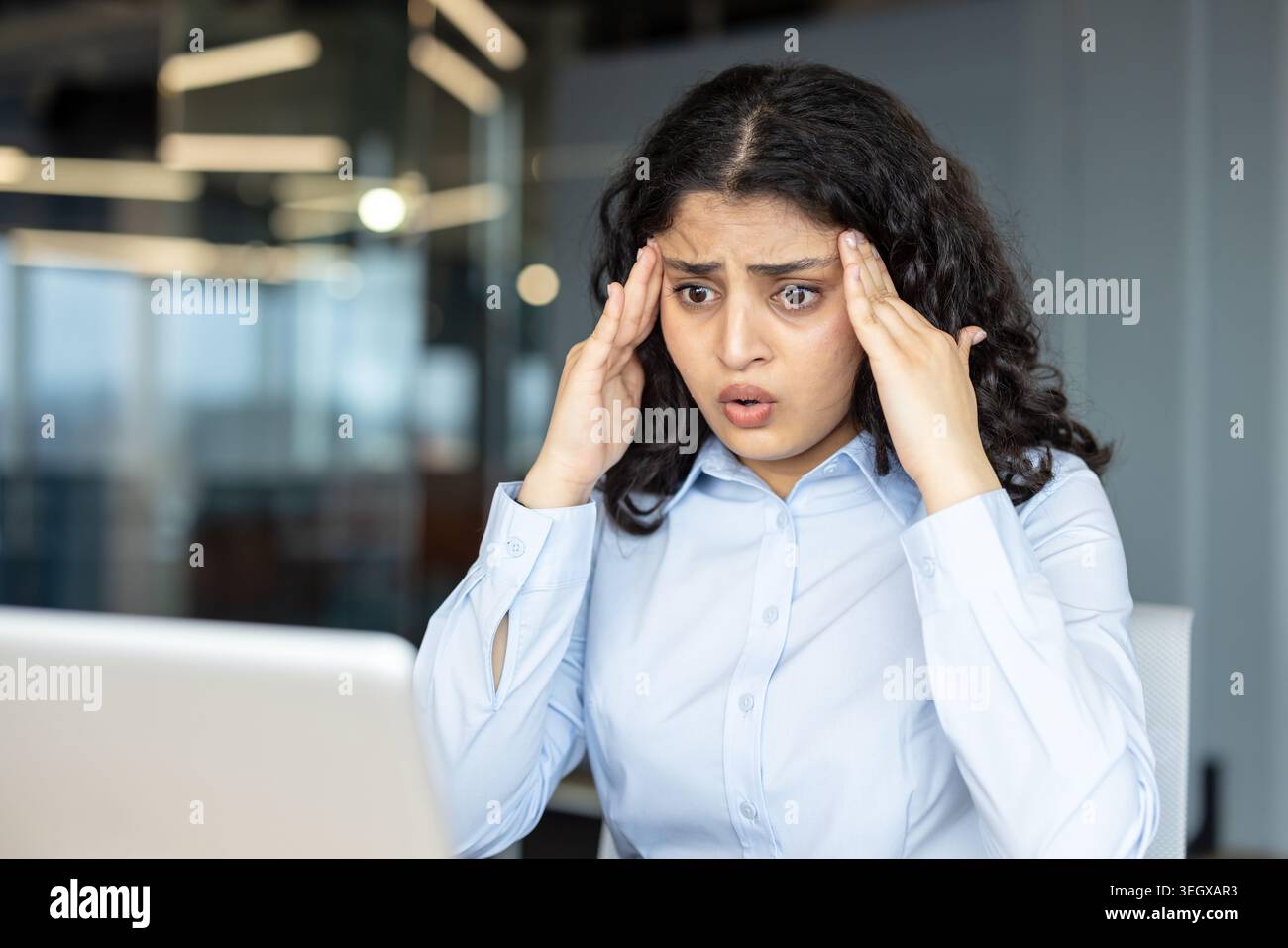 Giovane donna indiana che stringe la testa in un ufficio moderno, stressata e affaticata mentre lavora su un notebook, raffigurante ustione, ansia e pressione sul posto di lavoro Foto Stock