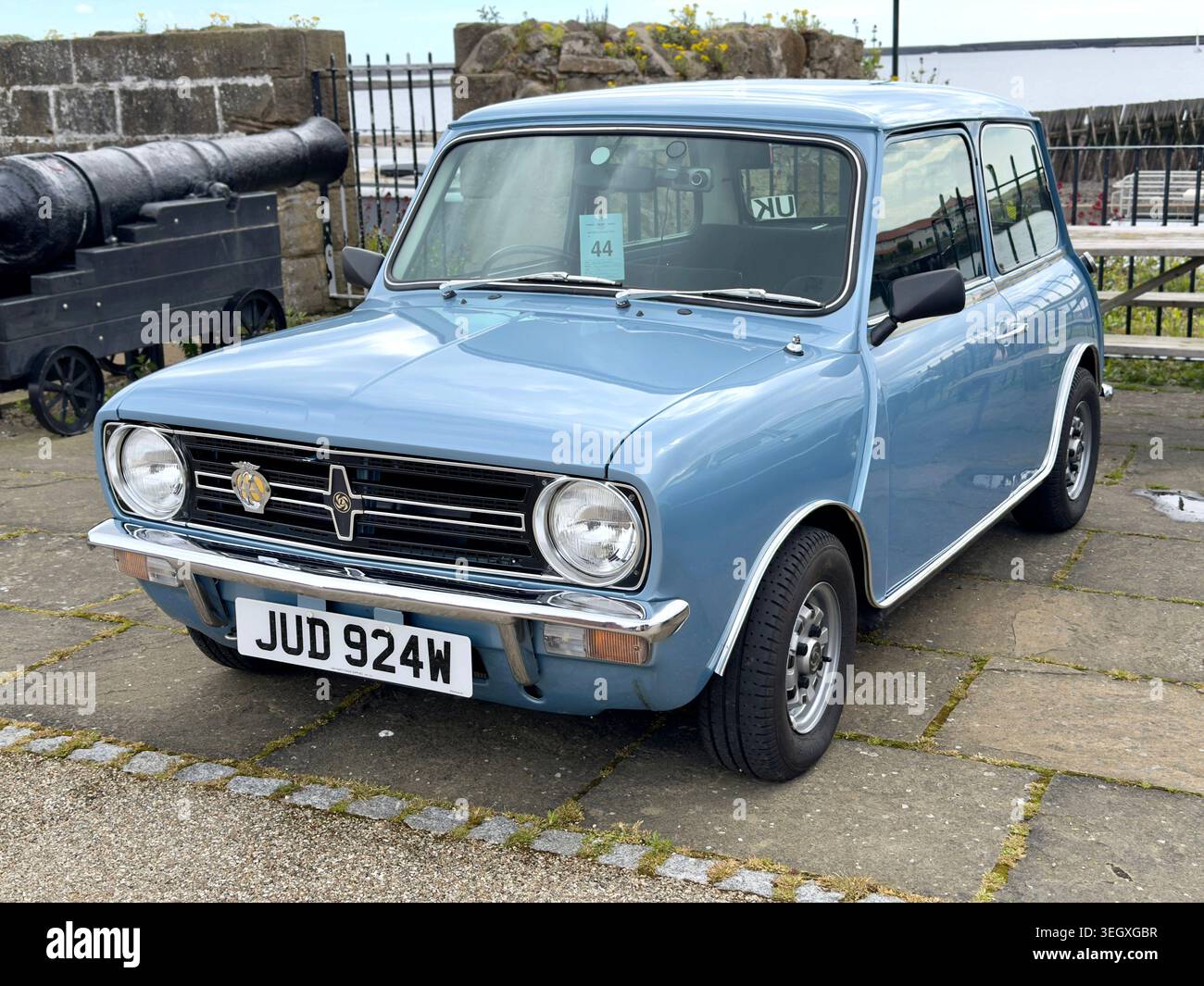 Classico Mini Clubman azzurro parcheggiato su un'area pavimentata con un cannone d'epoca sullo sfondo - Immagine stock catturata con smartphone