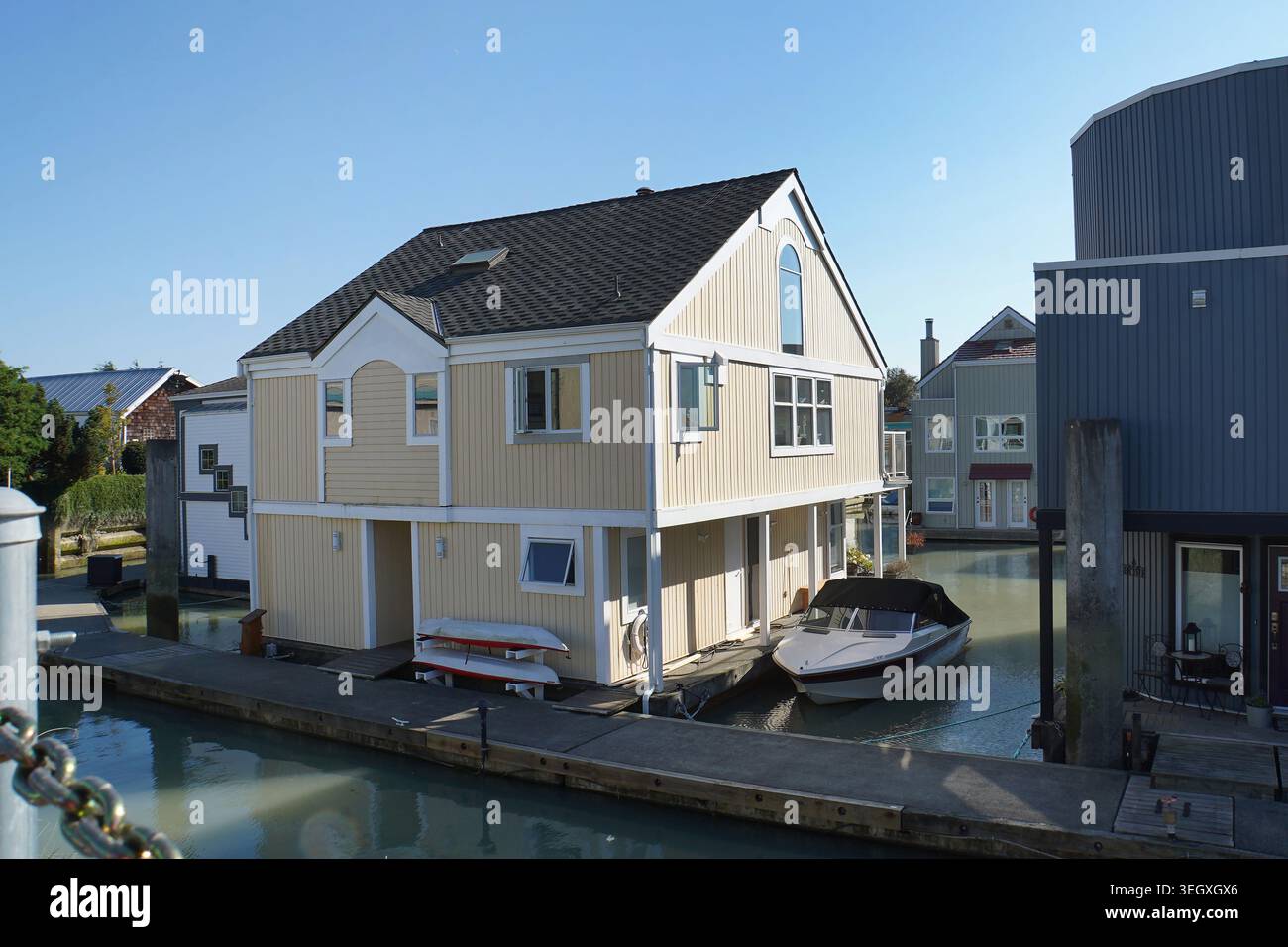Esterno di una moderna casa galleggiante a 2,5 piani di colore giallo pallido ancorata in una piccola comunità acquatica, accanto a un molo di legno sotto il cielo blu. Foto Stock