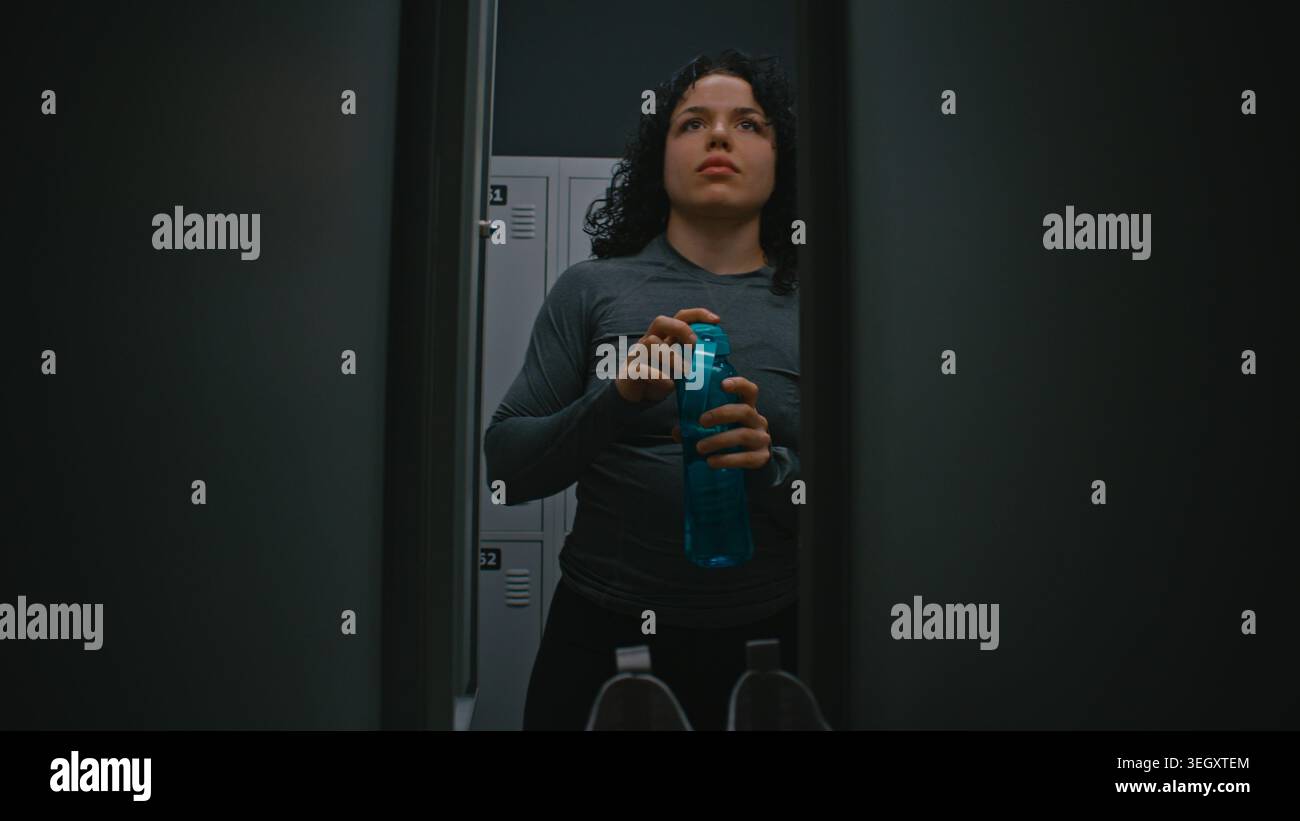 Atleta che si prepara per l'allenamento in Bright Locker Room. La donna cambia in Sportswear, beve acqua e chiude la porta dell'armadietto. Concetto di concentrazione, disciplina e inizio del viaggio atletico. Foto Stock