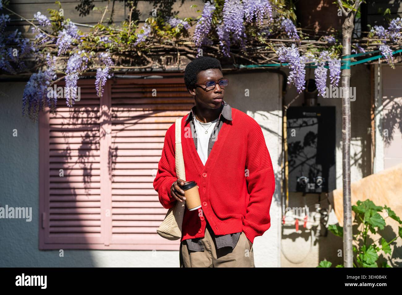 Studente afroamericano premuroso e bello in piedi sulla strada della città che regge una tazza di caffè. Foto Stock
