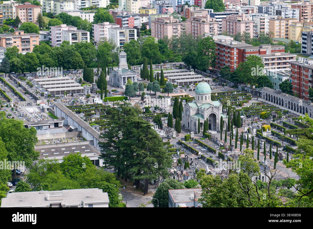 Vista aerea ad alto angolo del cimitero monumentale di Lugano, Svizzera. Chiesa storica con cupola verde, tombe simmetriche e cipressi Foto Stock