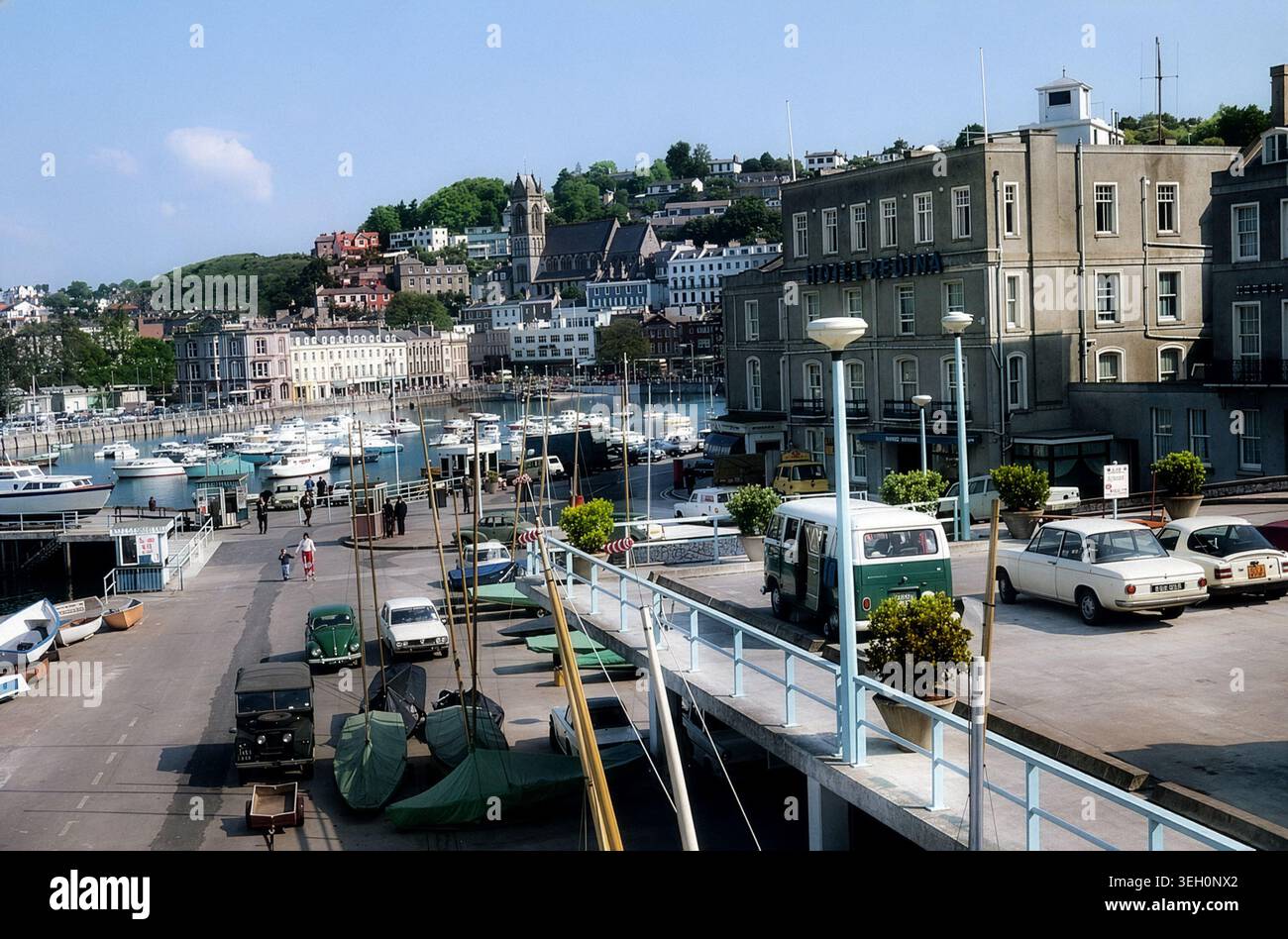 Torquay Harbour, Devon, Inghilterra, circa 1970. Vista sul porticciolo verso lo Strand e la chiesa di Santa Maria Maddalena, con l'Hotel Regina e una fila di auto d'epoca che costeggiano la banchina. La scena cattura la riviera inglese durante il boom del turismo post-bellico, prima di un'importante riqualificazione del lungomare e delle strutture del porto turistico. Foto Stock