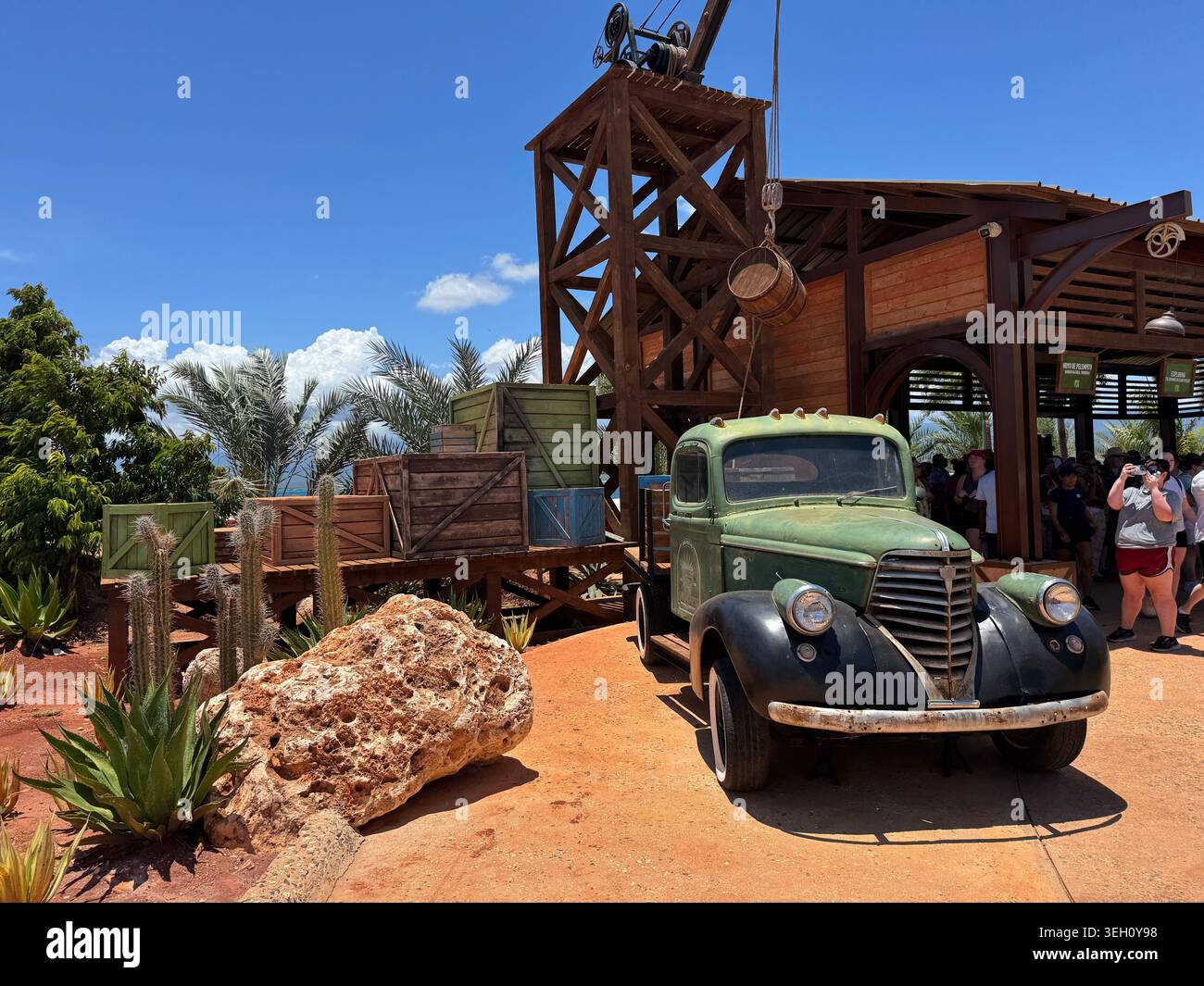 pick-up verde d'epoca parcheggiato in un parco a tema all'aperto con strutture minerarie rustiche in legno, rocce decorative e palme sotto il sole - Immagine stock catturata con smartphone