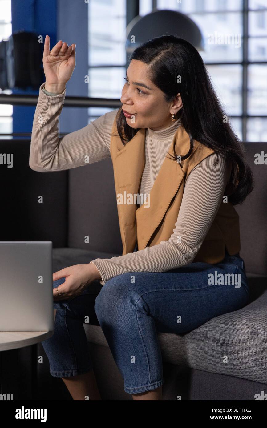Donna asiatica che indossa un gilet beige sopra il dolcevita, seduta sul divano in ufficio mentre gesti vicino al computer portatile Foto Stock