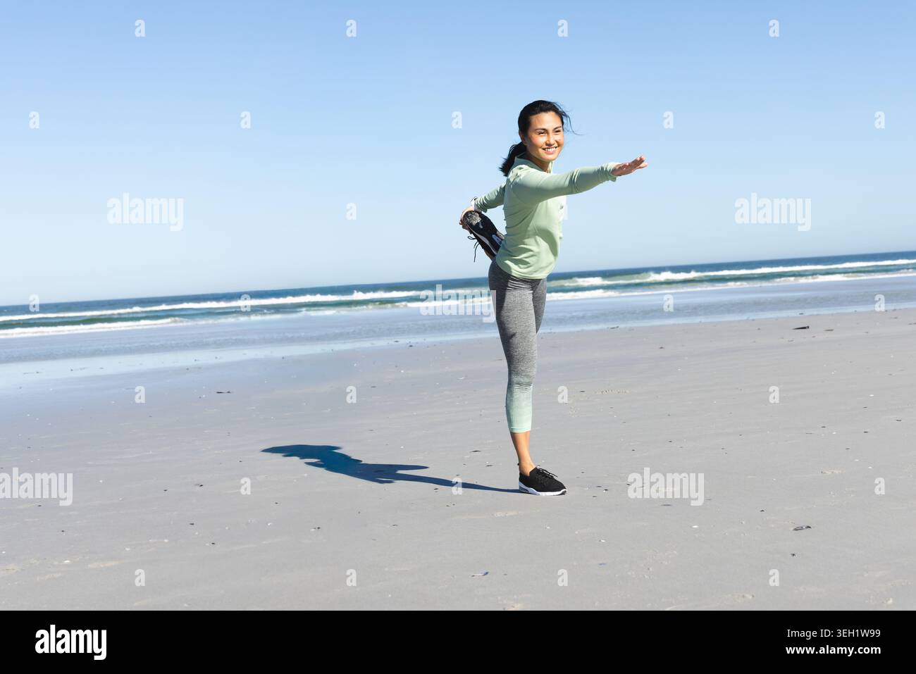 Donna asiatica degli anni '30 che si estende su una spiaggia di sabbia indossando leggings verdi che tengono la scarpa vicino alle onde Foto Stock