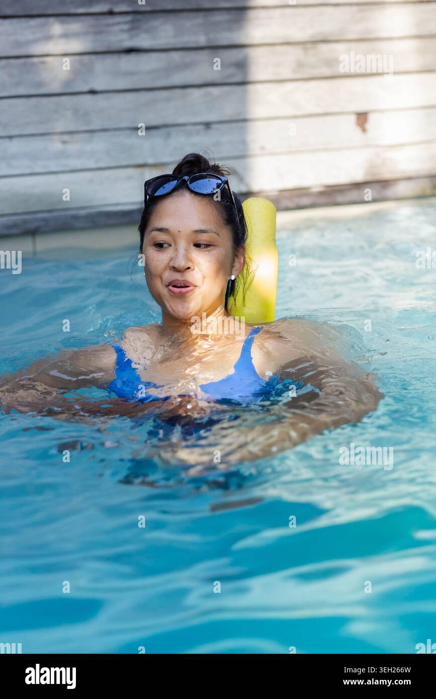 Donna che si rilassa, galleggia su una spaghetta gialla nella piscina sul retro con occhiali da sole e costume da bagno blu Foto Stock