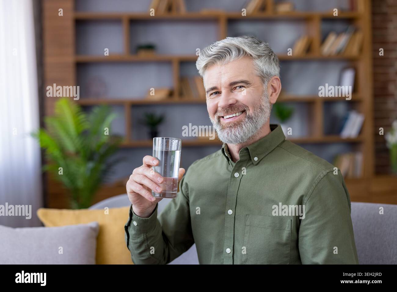Uomo maturo con una barba grigia e un sorriso genuino che promuove uno stile di vita sano mentre tiene un bicchiere d'acqua rinfrescante, nel suo ambiente confortevole Foto Stock