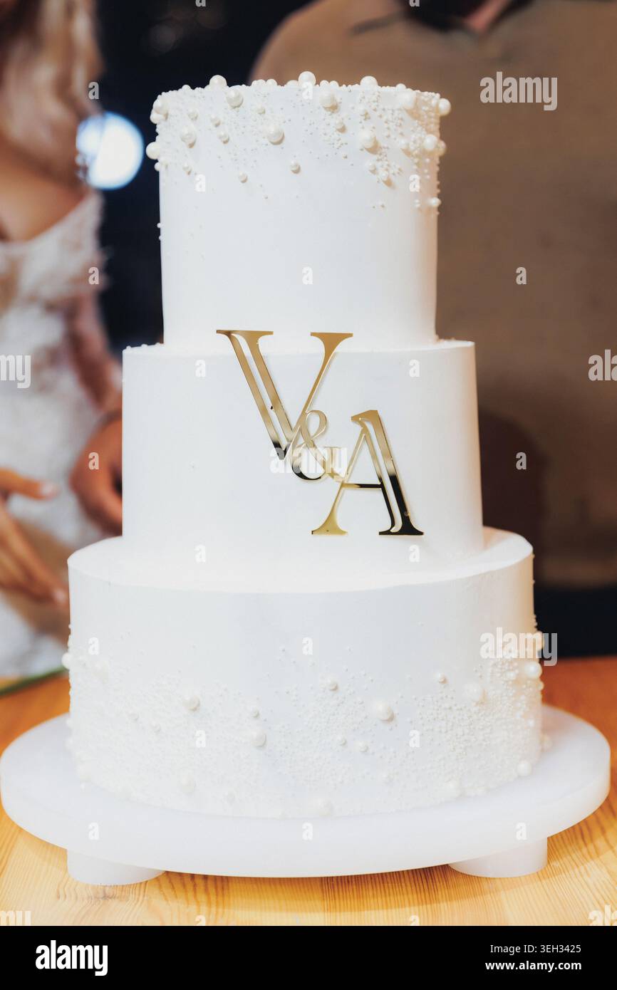Torta bianca a più piani decorata con un monogramma dorato e delicati dettagli perlati Foto Stock