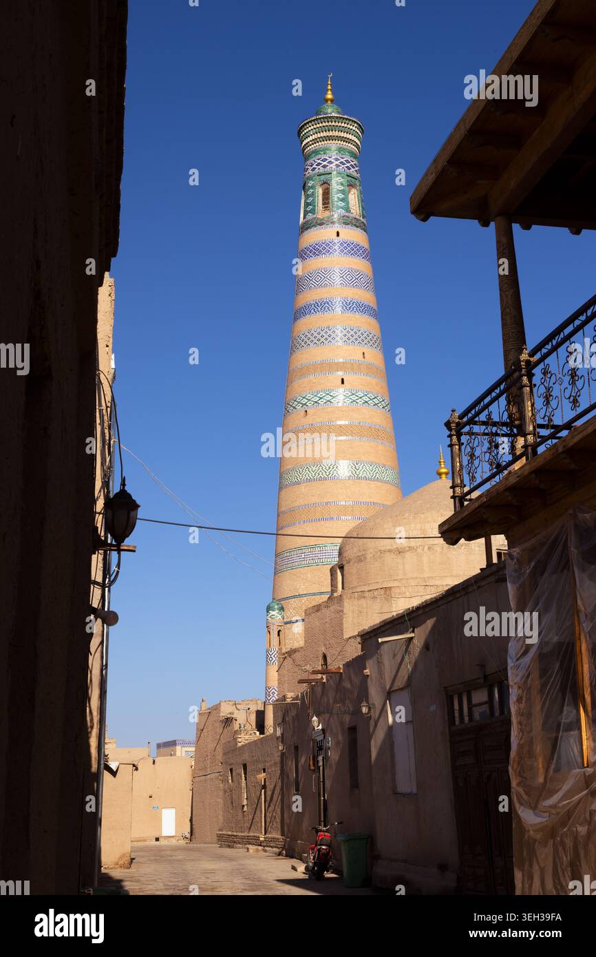 Islam Khoja Minaret visto attraverso uno stretto vicolo a Ichan Kala, Khiva. Fasce di piastrelle a righe e cupola verde si innalzano sopra le pareti di mattoni di fango sotto un blu chiaro Foto Stock
