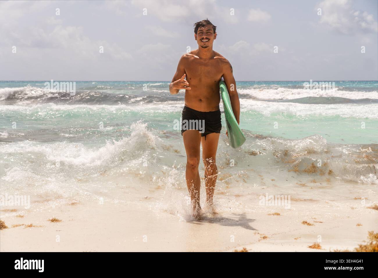 Giovane surfista che cammina fuori dall'oceano con i capelli bagnati, sorride e fa un gesto shaka mentre porta con sé una tavola da surf. Foto Stock
