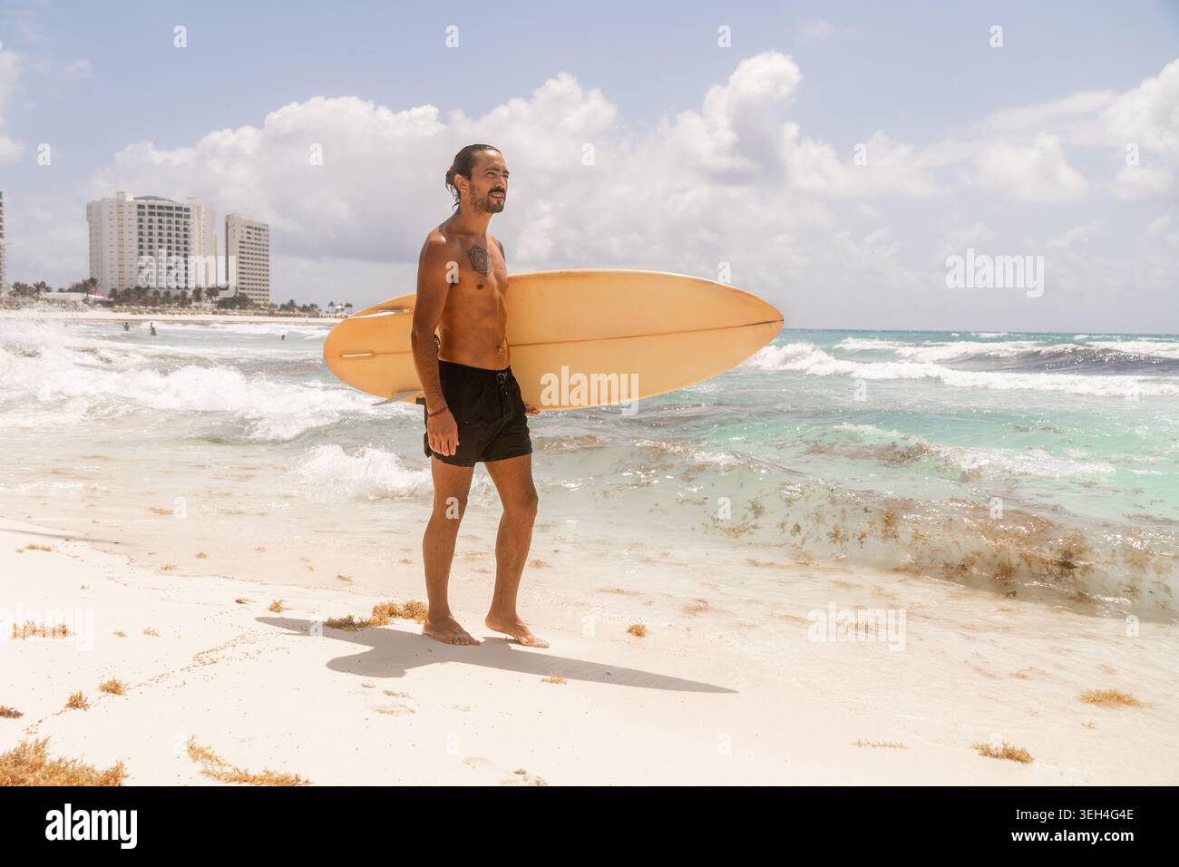 Indossa un surfista tatuato con lunghi capelli legati a un panino che porta una tavola da surf gialla vintage mentre cammini lungo una spiaggia luminosa. Foto Stock