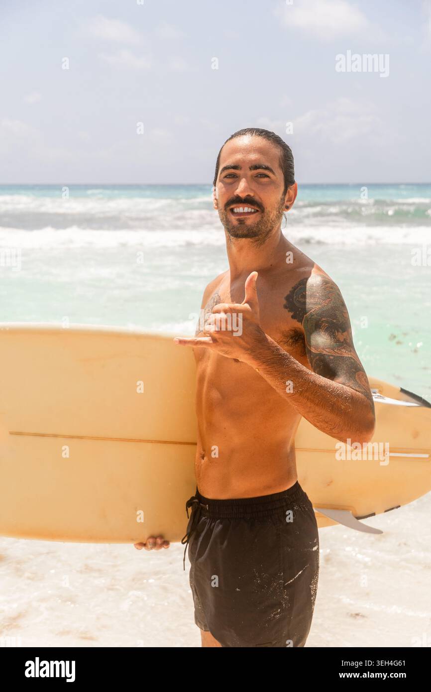Ritratto ravvicinato di un surfista sorridente con tatuaggi e un panino che fa un gesto shaka tenendo in mano una tavola da surf gialla d'epoca. Foto Stock