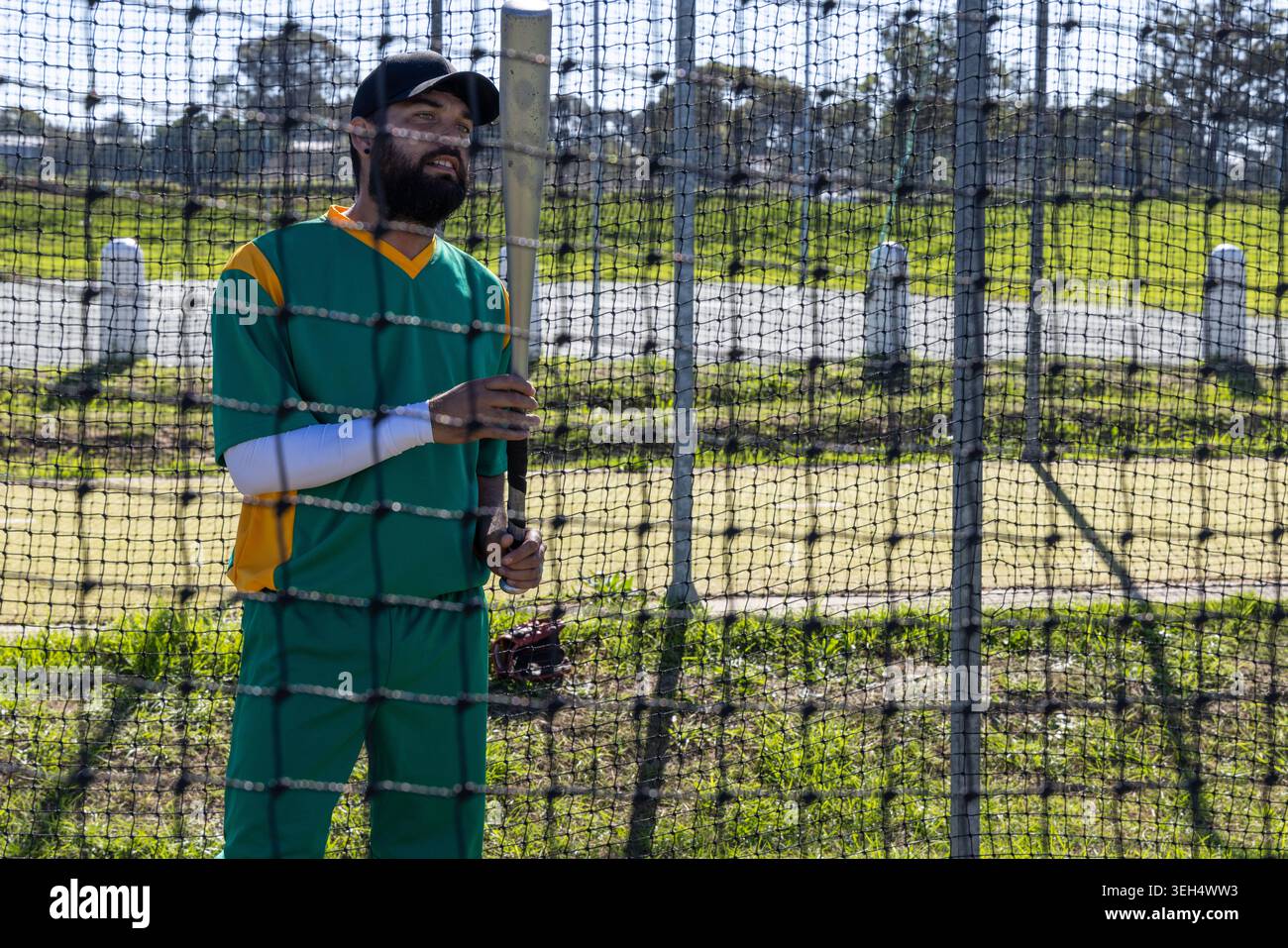 Uomo medio-adulto che indossa un'uniforme verde-gialla in piedi dietro una rete che tiene una mazza di legno nel parco Foto Stock