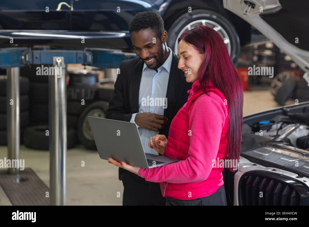 Donna meccanica in piedi accanto a un'auto con cofano aperto, che spiega l'elenco di controllo per la manutenzione del veicolo a un cliente utilizzando un notebook. Foto Stock