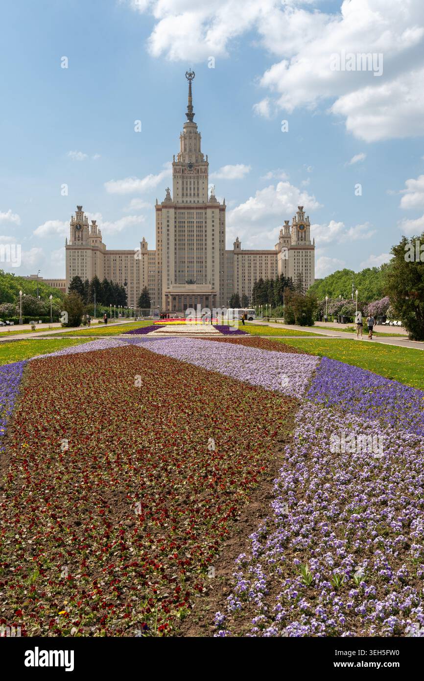 Mosca, Russia - 17 maggio 2026: Lunghe aiuole di fiori conducono verso l'edificio principale dell'Università statale di Mosca con fontane, vicoli del parco e spettacolari nuvole primaverili Foto Stock