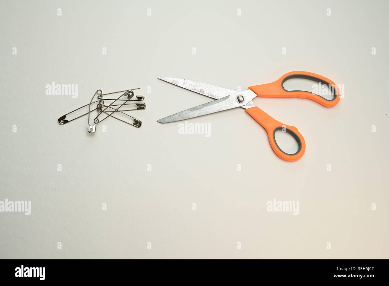 Forbici con manici arancioni e spille di sicurezza in metallo disposte su uno sfondo semplice, una natura morta minimalista di strumenti per il cucito e di fissaggio domestico. Foto Stock