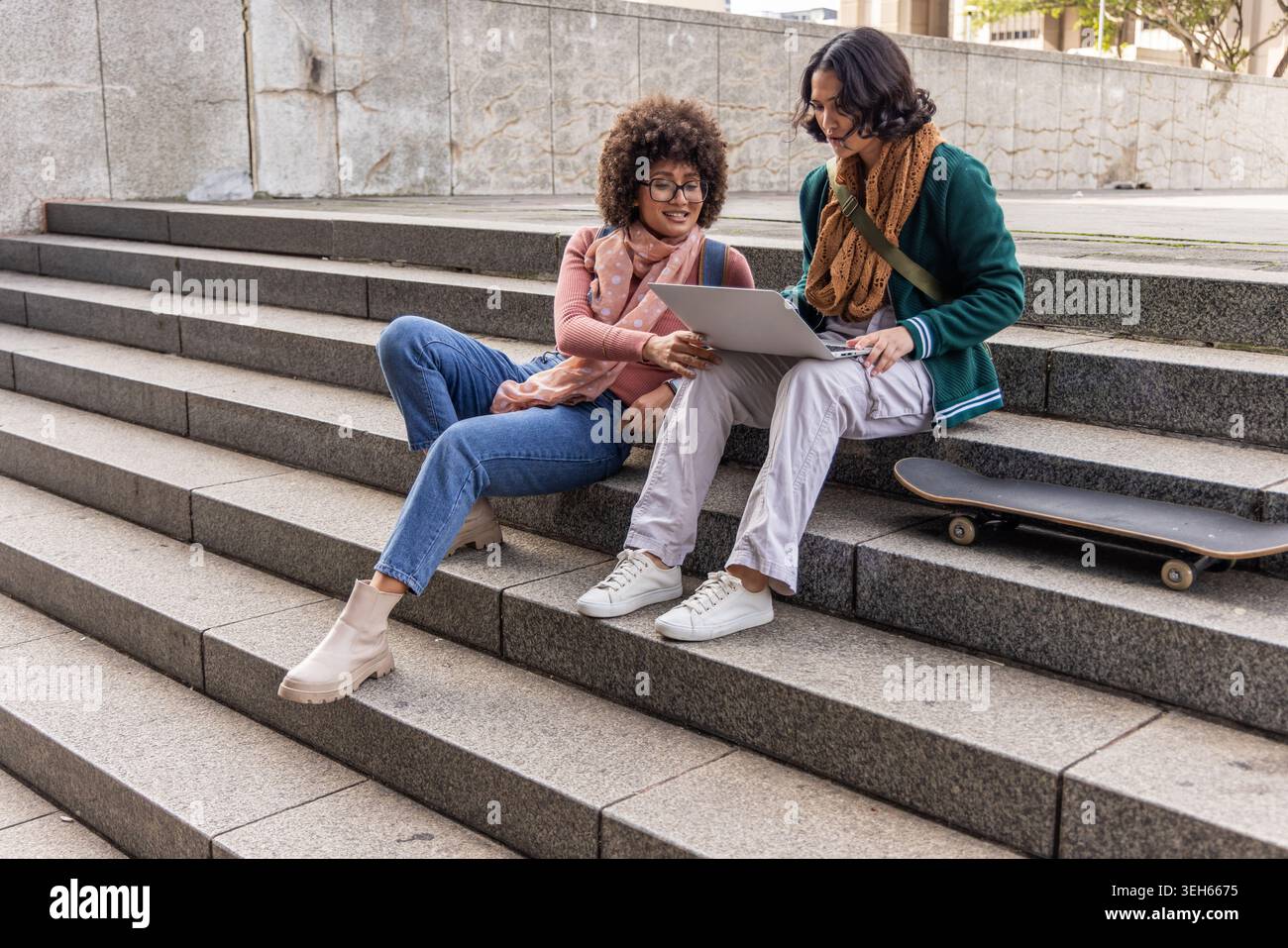 Diverse studentesse che studiano insieme sui gradini del campus, condividendo laptop con skateboard nelle vicinanze Foto Stock
