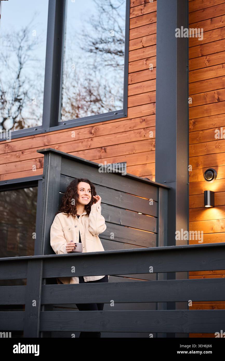 Donna sorridente che gode dell'aria fresca sulla terrazza di una cabina elegante, ideale per concetti di benessere, tempo libero, vita remota e fascino immobiliare Foto Stock