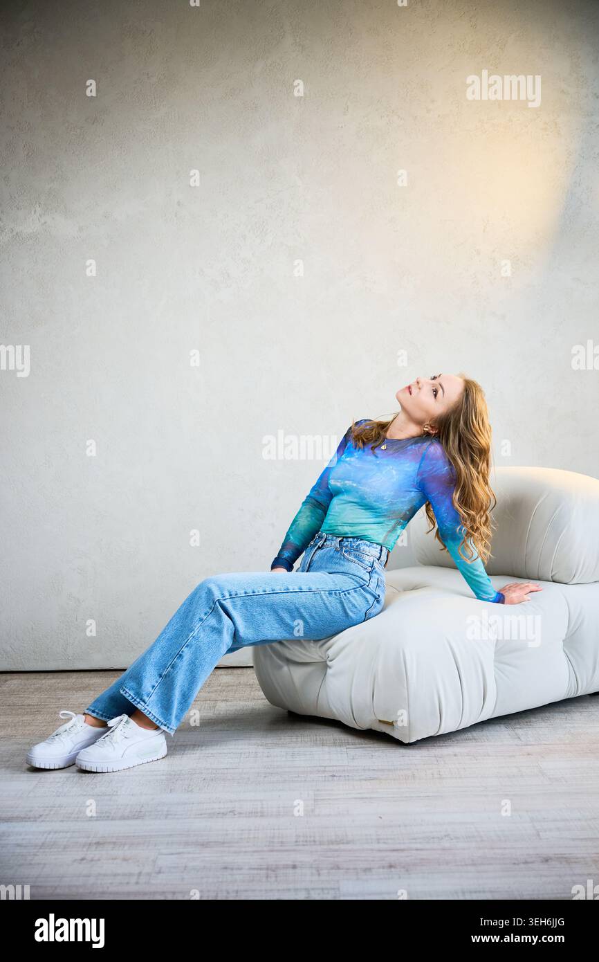 Una giovane donna si appoggia su una lussuosa sedia, gli occhi chiusi, assaporando un'atmosfera tranquilla in uno spazio arioso e luminoso. I colori tenui e la luce naturale creano un'atmosfera serena per il relax. Foto Stock