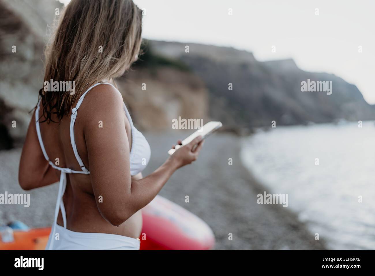 Donna sulla spiaggia dello smartphone. Giovane donna che si rilassa su una spiaggia di ciottoli usando il suo telefono vicino alle scogliere. Foto Stock