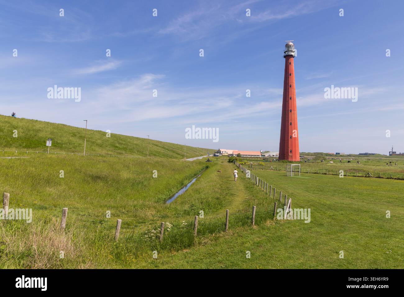 Den Helder, Paesi Bassi. Un'ampia vista panoramica dello storico faro rosso di Lange Jaap, che si erge su un cielo blu, circondato da dighe verdi erbose Foto Stock