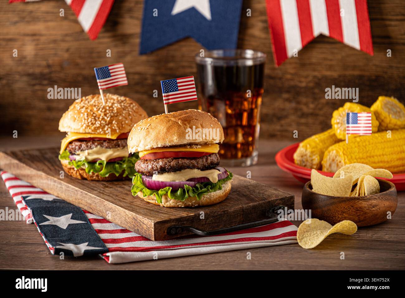 Cheeseburger fatti in casa con bandiere americane, cola corn e patatine fritte su un rustico tavolo di legno per festeggiare il 4 luglio e festeggiare il barbecue estivo. Foto Stock
