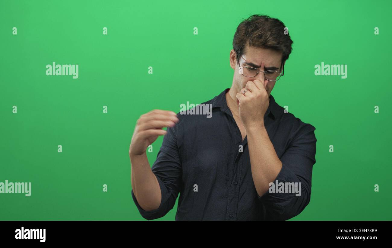 Uomo che pizzica il naso con le dita mentre alza l'altra mano, indossa occhiali e una camicia nera contro uno studio di green screen; disgusto. Foto Stock