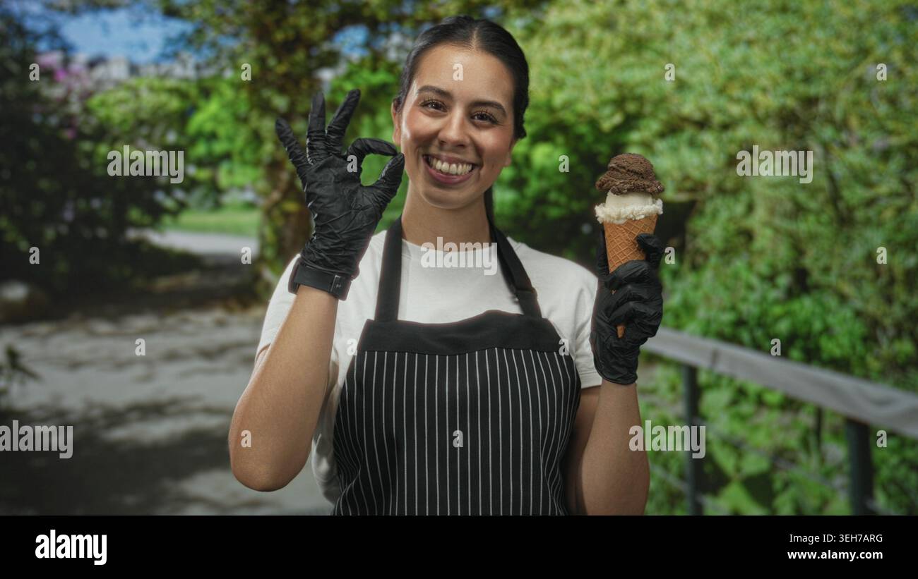 Donna in grembiule che tiene il cono del gelato e fa un gesto discreto nella foresta, sorride alla macchina fotografica; servizio allegro. Foto Stock