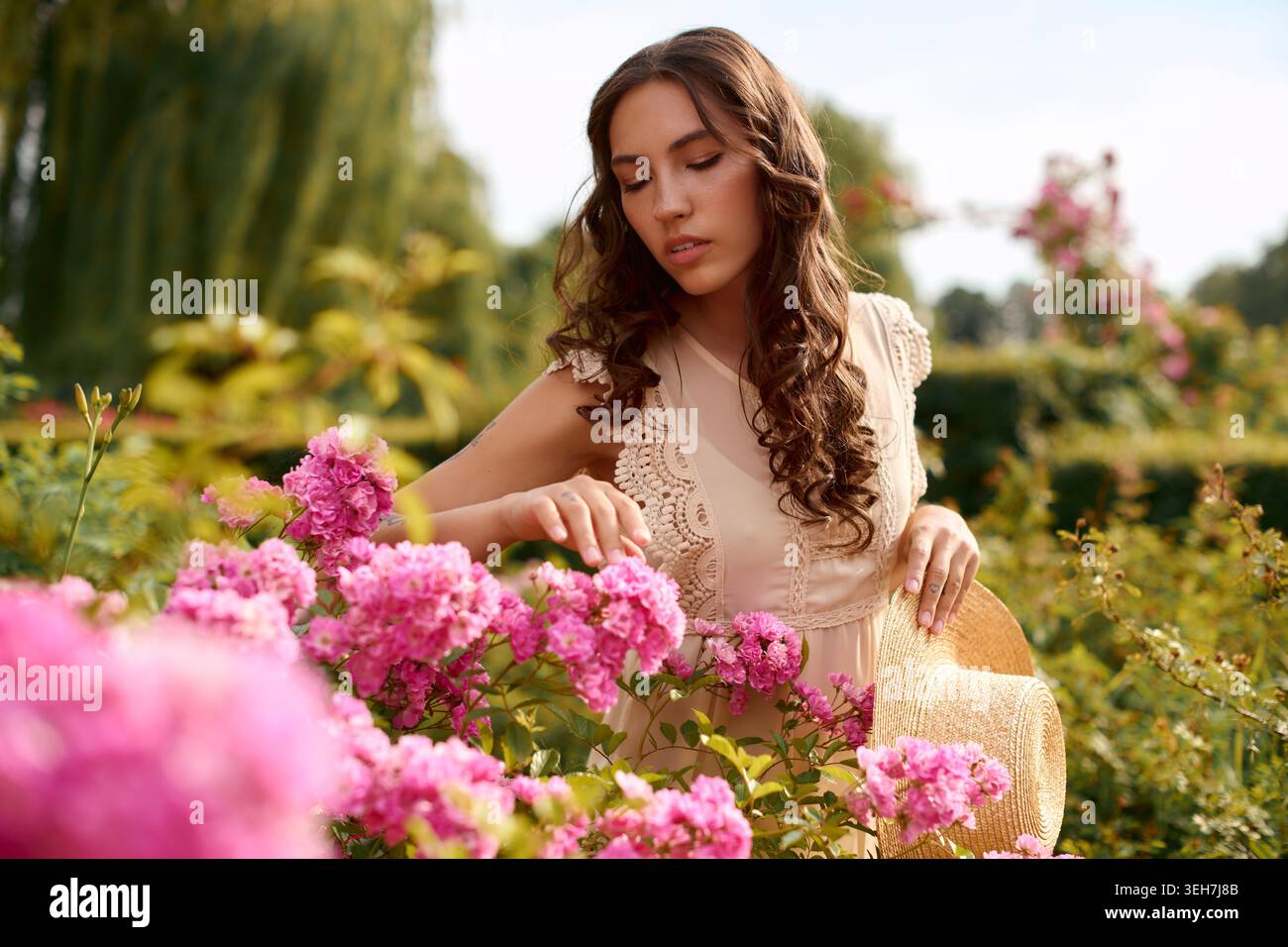 Una donna adorabile tocca delicatamente i fiori rosa circondati dal verde in un parco tranquillo. Foto Stock