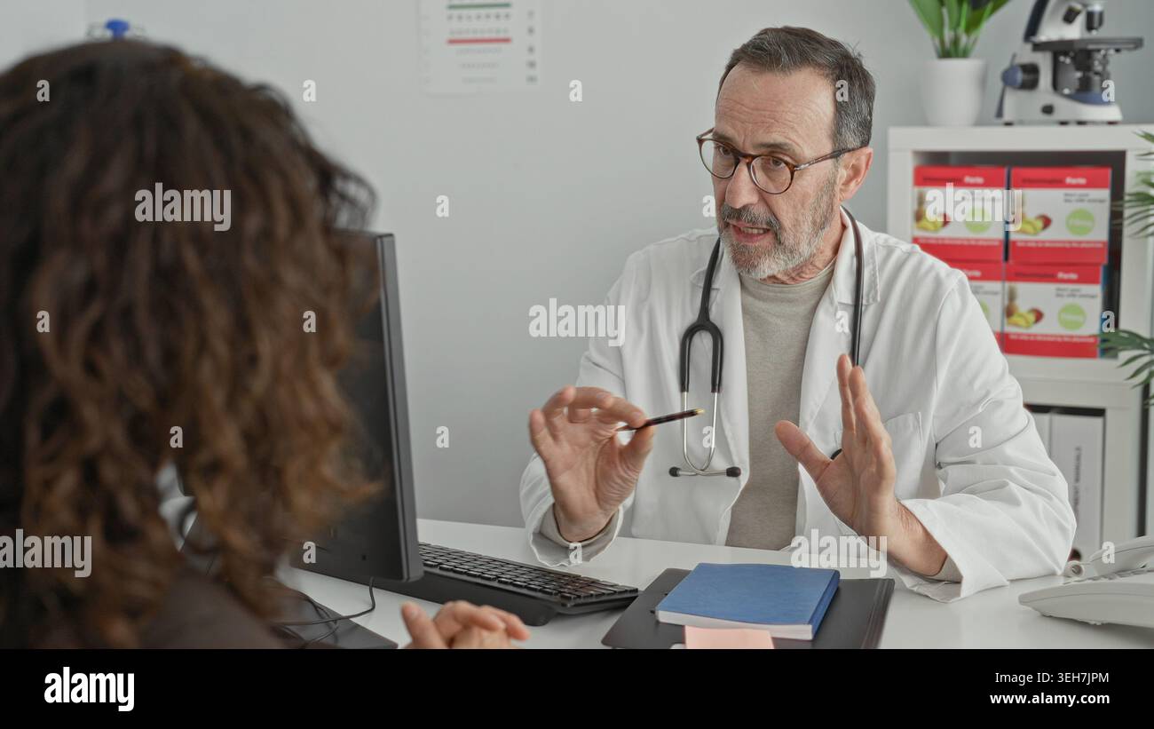 Uomo di mezza età medico con stetoscopio tiene la penna e i gesti mentre consulta il paziente alla scrivania con computer e cartella medica nell'edificio della clinica; re Foto Stock
